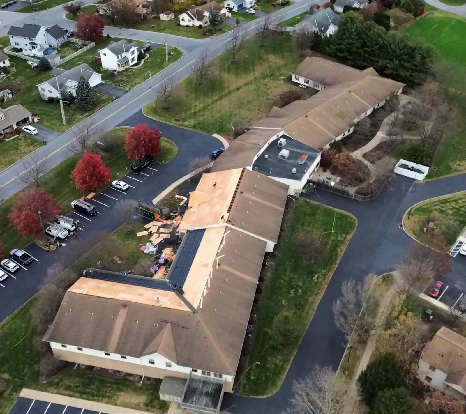 Aerial of a long multi-family building with the old roof partly torn off, bare decking and new shingles on different sections and debris staged by the parking lot